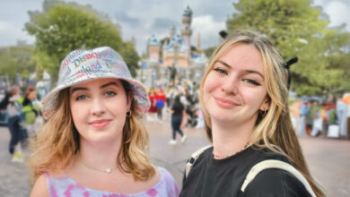 Two young women smiling at the camera in front of Sleeping Beauty Castle at Disneyland. One wears a pastel tie-dye tank top and a bucket hat with “Disneyland” printed across it, while the other wears a black shirt with winged eyeliner and light blonde hair. The castle and park guests are softly blurred in the background.